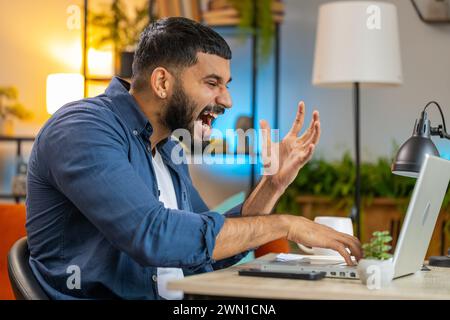 Angry furious Indian man freelancer using laptop while working at home office having nervous breakdown at work, stress management, mental distress problems, losing temper, reaction on failure. Stock Photo