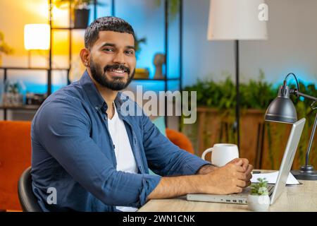 Portrait of smiling young bearded male freelancer in casual clothes sitting at table with laptop working remotely at home office. Confident Indian Hispanic businessman happily looking at camera. Stock Photo