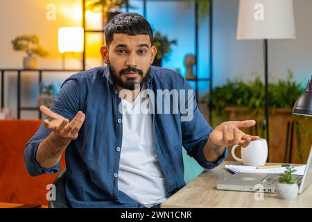 I don't know. Confused Indian bearded man working on laptop at home office workplace. Freelancer feeling embarrassed about ambiguous question, having doubts, no answer idea, being clueless, uncertain. Stock Photo
