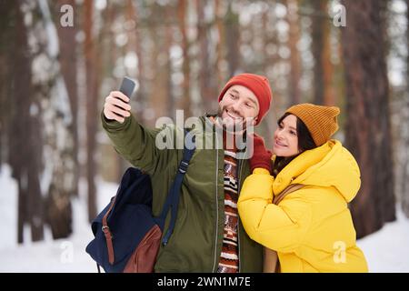 Young Caucasian man and woman in love taking selfie on smartphone while walking in forest on cold winter day Stock Photo