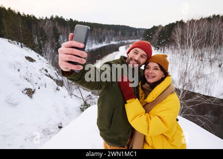 Young Caucasian man and woman in love taking selfie on smartphone while spending winter day hiking in mountains Stock Photo