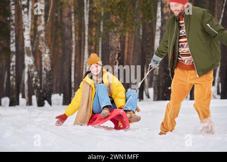 Bearded man having fun on cold winter day pulling sledge with his girlfriend sitting on it Stock Photo