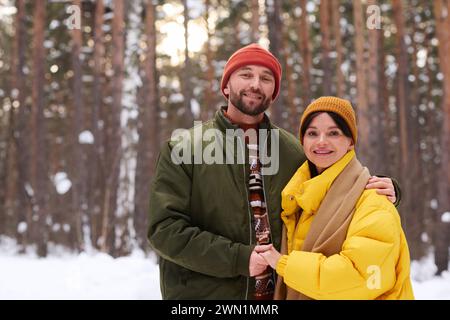 Medium portrait of young couple smiling at camera while standing in forest on sunny winter day Stock Photo