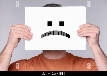 Man hiding behind sheet of paper with sad face on grey background Stock Photo