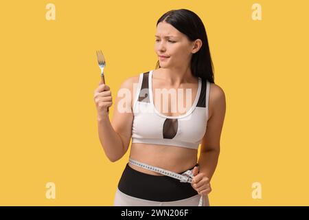 Beautiful young displeased woman with fork and measuring tape on yellow background. Weight loss concept Stock Photo