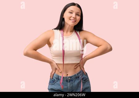 Beautiful young happy woman in loose jeans with measuring tape on pink background. Weight loss concept Stock Photo