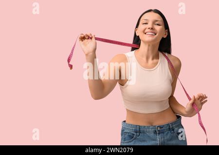 Beautiful young happy woman in loose jeans with measuring tape on pink background. Weight loss concept Stock Photo