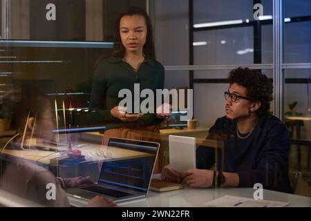 Young female programmer standing at table having discussion with her colleagues in modern office in evening Stock Photo