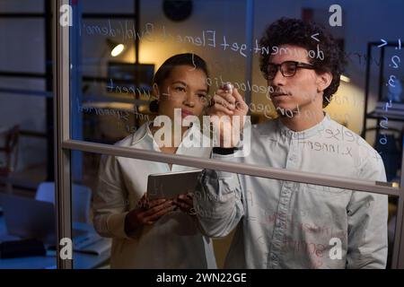 Young man and woman working together on code writing it on glass wall in IT company office late in evening Stock Photo