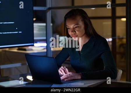 Medium shot of young woman developing code using laptop in office late at night Stock Photo