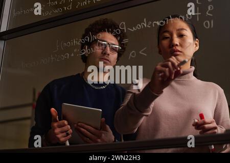 Ethnically diverse male and female programmers working together on code writing it on glass wall in office late in evening Stock Photo