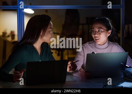 Two young ethnically diverse female programmers working together on code using laptops in office late in evening Stock Photo