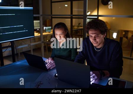High angle view of ethnically diverse young programmers coding on laptops at workplace in IT company office late in evening Stock Photo