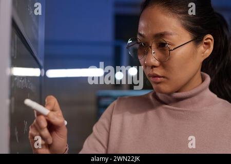 Medium closeup of young Asian programmer working on code in office late in evening, writing it on glass wall Stock Photo