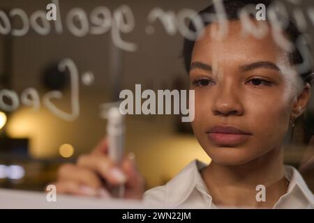 Closeup of young biracial female programmer writing code on glass wall in office late in evening Stock Photo