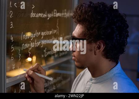 Side view closeup of young man developing code writing it on glass wall in office late at night Stock Photo