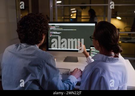 Rear view shot of young man and woman fixing computer code in office late in evening Stock Photo