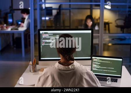 High angle rear view of unrecognizable young female programmer working on code using desktop computer and laptop in office at night Stock Photo