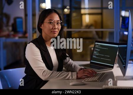Medium portrait of young Asian female programmer working on code using laptop and desktop computer at workplace in IT company office late in evening Stock Photo