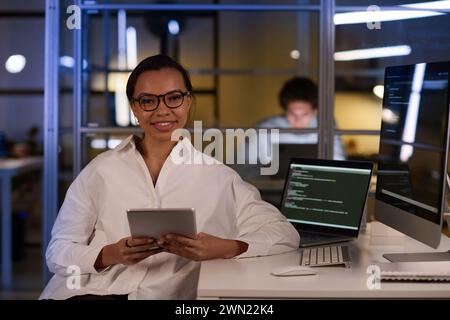 Medium portrait of young woman wearing eyeglasses holding digital tablet sitting at workplace in office late in evening Stock Photo