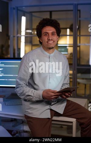 Vertical medium portrait of young male programmer sitting on desk in office late in evening Stock Photo