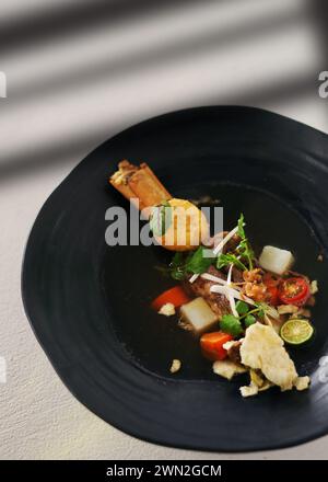A black bowl filled with vegetables and broth being poured from a ladle on a wooden table with white tablecloth Stock Photo