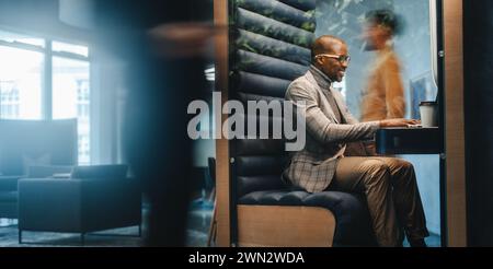 Young man sitting in a busy coworking office, working on a laptop inside a booth. Business people are moving around in the shared workspace. Professio Stock Photo