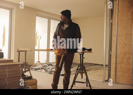 Smiling builder stands in a kitchen, wearing a tool belt, doing home renovation. He is focused on upgrading the flooring with various equipment, showc Stock Photo