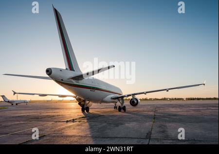 Rear three quarter view of passenger airplane on airport apron and few other planes in the background. Low evening sun creates a long shadow. Stock Photo