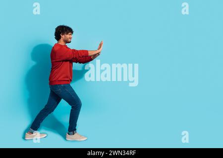 Full length profile portrait of hardworking young man arms push heavy empty space isolated on blue color background Stock Photo
