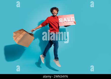 Full length size photo of overjoyed curls handsome shopaholic hold red banner sale and eco paper bags isolated on blue color background Stock Photo