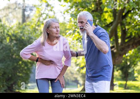 Elderly woman feeling abdominal pain while exercising, concerned man with beard making emergency call in park. Stock Photo