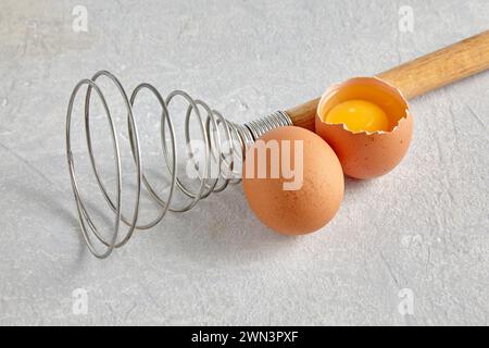Two beige hen's eggs with yolks and a spiral whisk to whip on a gray concrete countertop Stock Photo