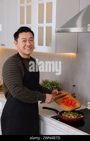 Cooking process. Happy man adding cut bell pepper into frying pan in kitchen Stock Photo