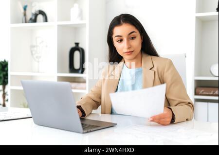 Work with documents. Focused serious indian or arabian business woman, financial manager, sit at workplace in the office with laptop, concentrated studying documents, create business plan and strategy Stock Photo