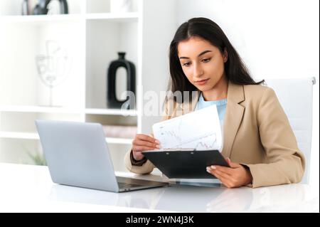 Concentrated serious indian or arabian business woman, financial manager, sitting at workplace in the office, using laptop, doing paperwork, focused studying documents, create business plan Stock Photo