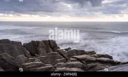 Sunset over rough ocean coast with waves and pancake like rocks in foreground, New Zealand Stock Photo