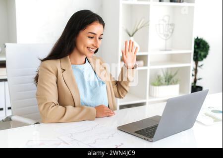 Online conference. Smiling successful indian or arabian business lady, company team leader, sits in a modern office, using laptop for online video conference with business team or clients, greeting Stock Photo
