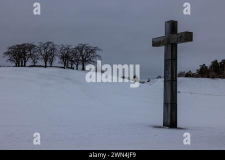 snowy landscape of stockholm woodland cemetery large granite cross at the foot of a small hill topped with dark elm trees skogskyrkogarden Stock Photo