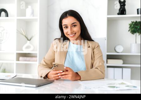 Beautiful friendly arabian or indian beautiful business woman, manager, ceo, sits at work desk in the office, using mobile phone, chatting with partners or customers, typing message, smile at camera Stock Photo