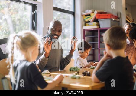 Smiling male teacher holding clay while sitting with kids in classroom at preschool Stock Photo