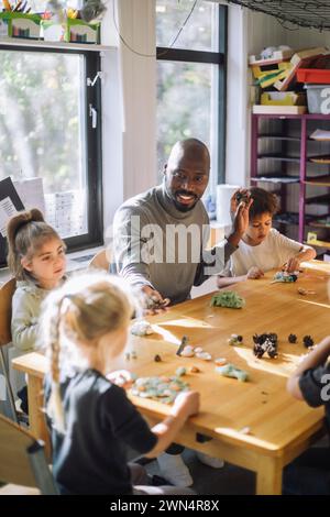 Male teacher playing with kids while sitting at bench during art class at preschool Stock Photo