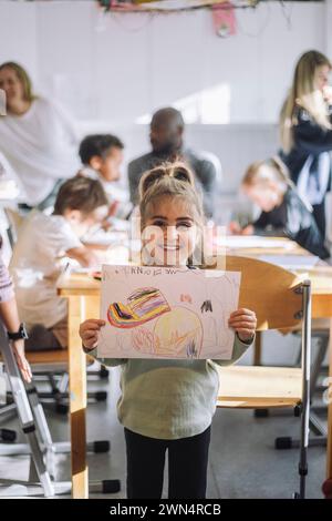 Portrait of smiling girl showing drawing while standing near bench in classroom at kindergarten Stock Photo