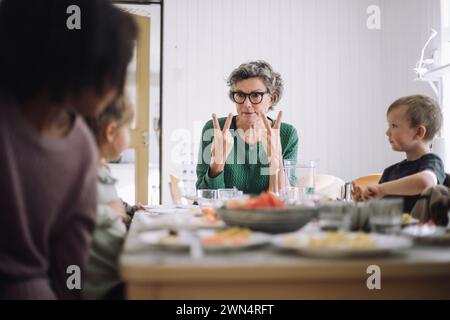 Senior female teacher communicating with students through hand signs while sitting at dining table during lunch break Stock Photo