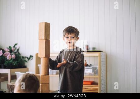 Boy looking away while standing near tower of toy blocks in classroom at kindergarten Stock Photo