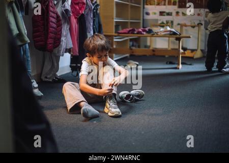 Boy tying shoelace while sitting on floor in cloakroom at preschool Stock Photo
