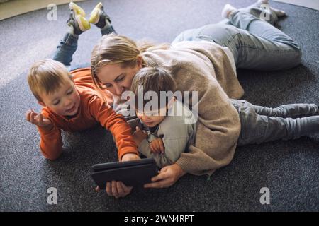 High angle view of kids watching digital tablet with female teacher lying down on carpet at preschool Stock Photo