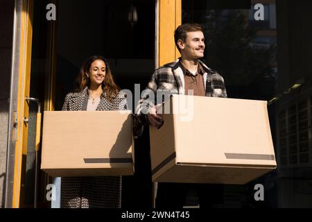 Smiling young couple carrying boxes while walking outside apartment building Stock Photo