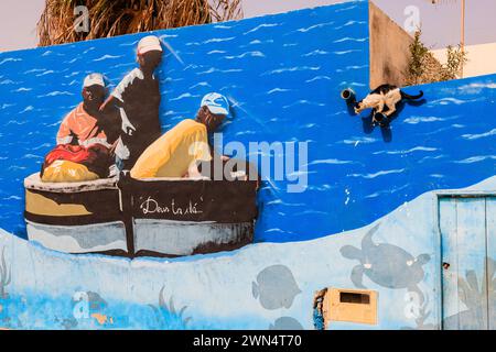 a cat perched on a wall appears to be preparing to jump into a fishing boat which is really a brightly painted mural on a wall in palmeira cape verde Stock Photo