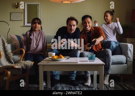 Excited gay couple competing while playing video game with daughter sitting on sofa at home Stock Photo